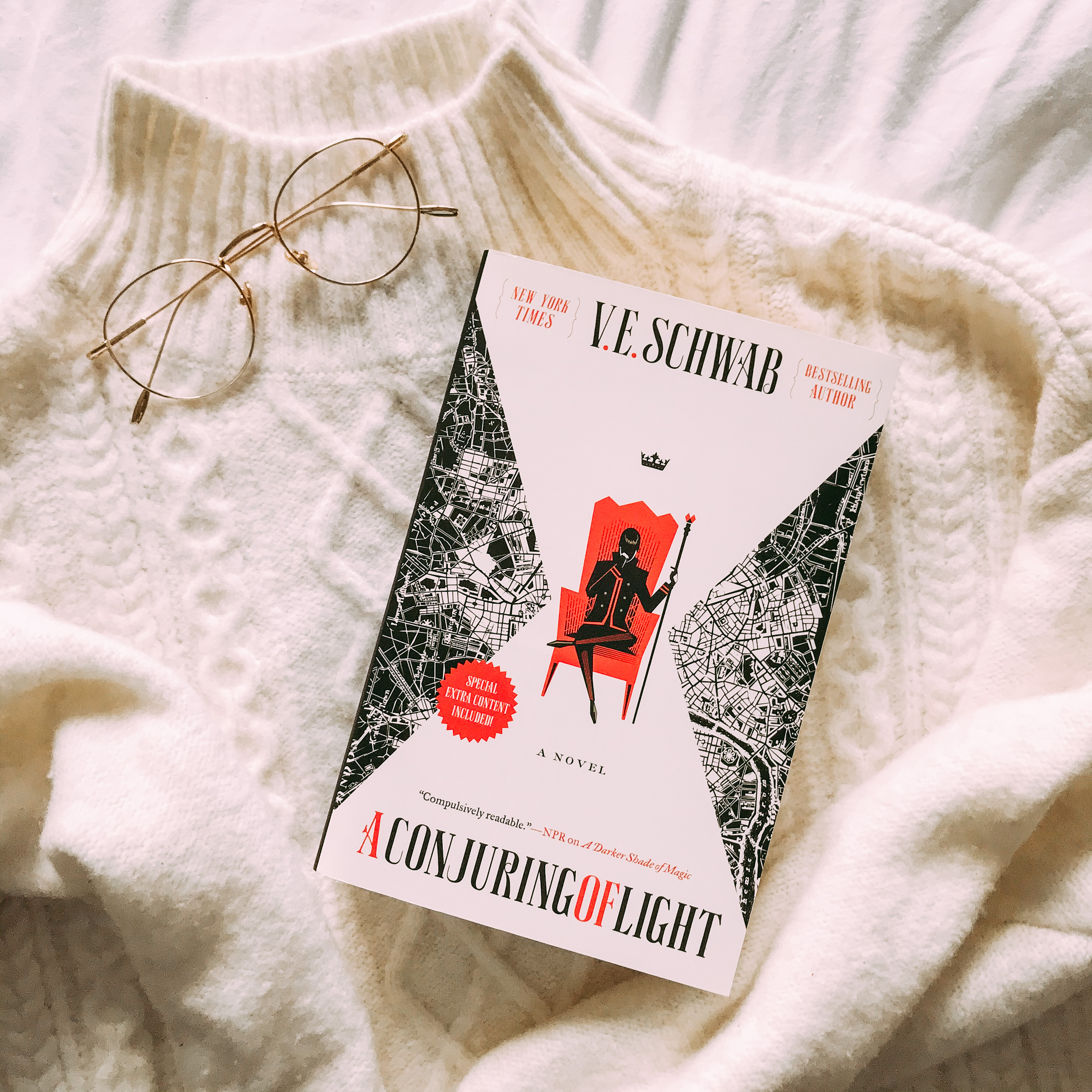 a conjuring of light book by ve schwab with a white turtleneck sweater and gold rimmed glasses